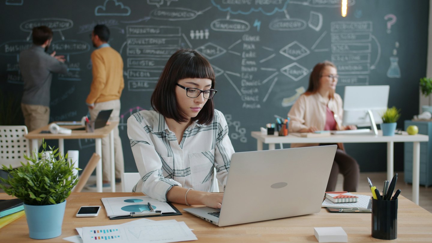 Kobieta w okularach pracuje przy laptopie przy drewnianym biurku.